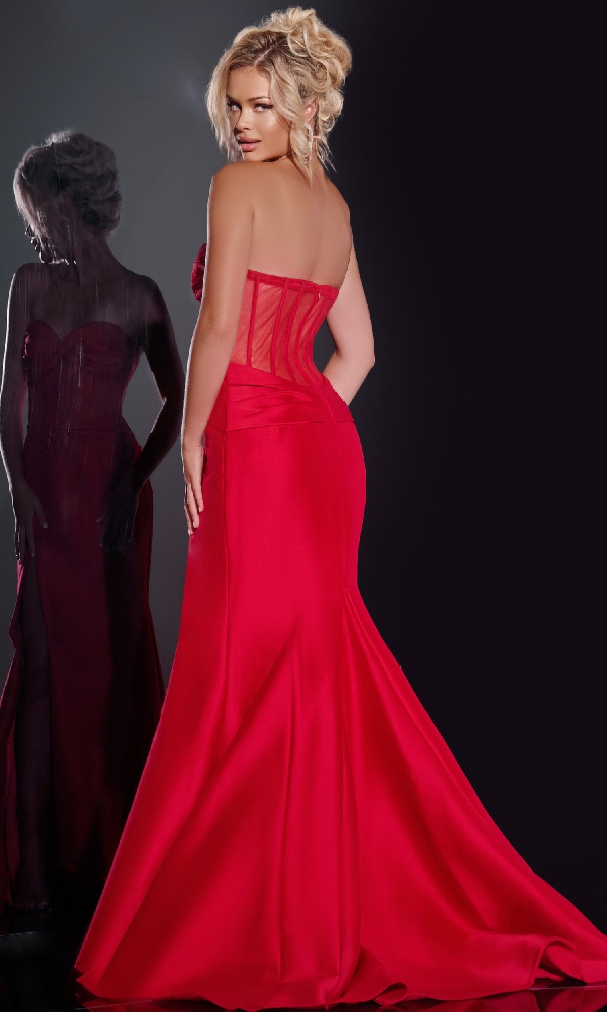 A woman wearing the Jovani 42375 long prom dress, featuring a strapless corset bodice, stands with her back to the camera and looks over her shoulder as her reflection glimmers in a mirror.